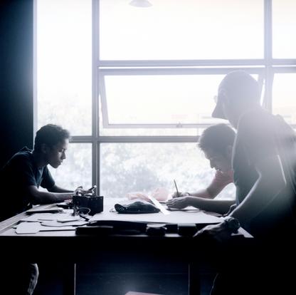 Three men writing at a table backlit by the open window behind them