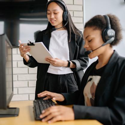 Two women talking wearing headsets, one is typing on a computer in the foreground and one is holding paper next to her