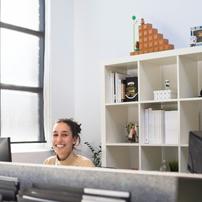A woman in a cubicle smiling at the camera over a desktop monitor