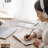 A woman wearing headphones writing in a composition notebook in front of her laptop