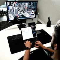 A man typing on a laptop in front of a computer monitor displaying geometric patterns