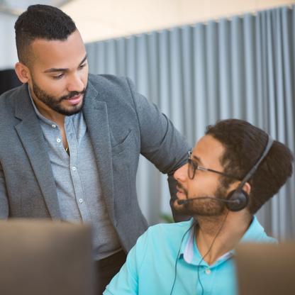 A man with a headset sitting a t a computer talking to another man over his shoulder