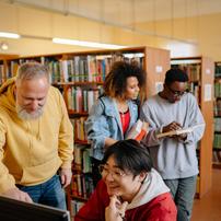Two people looking in a book in the background and two people looking at a computer in the front in a library