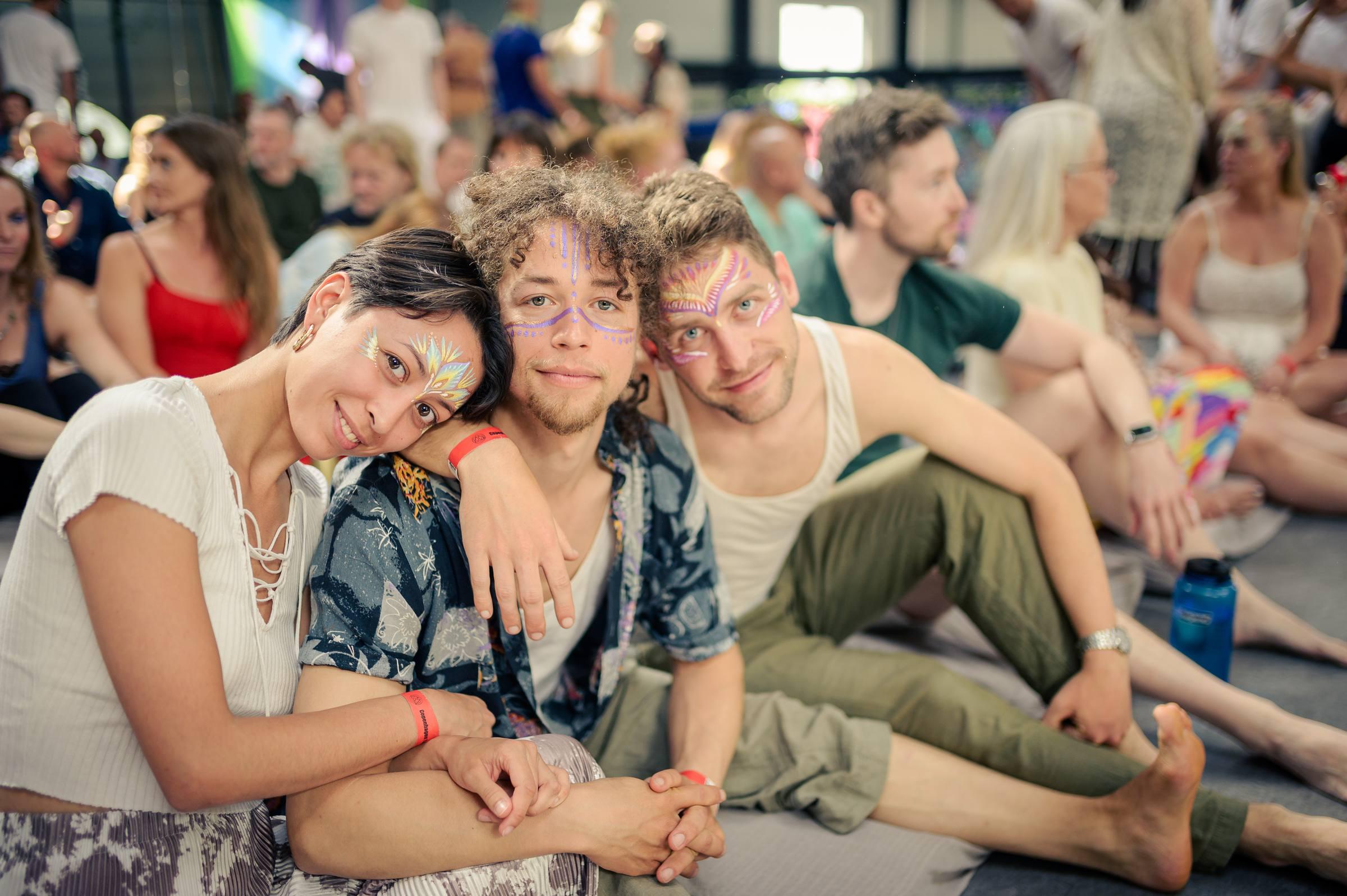 Three smiling participants with festival face paint sitting close together at Copenhagen Tantra Festival