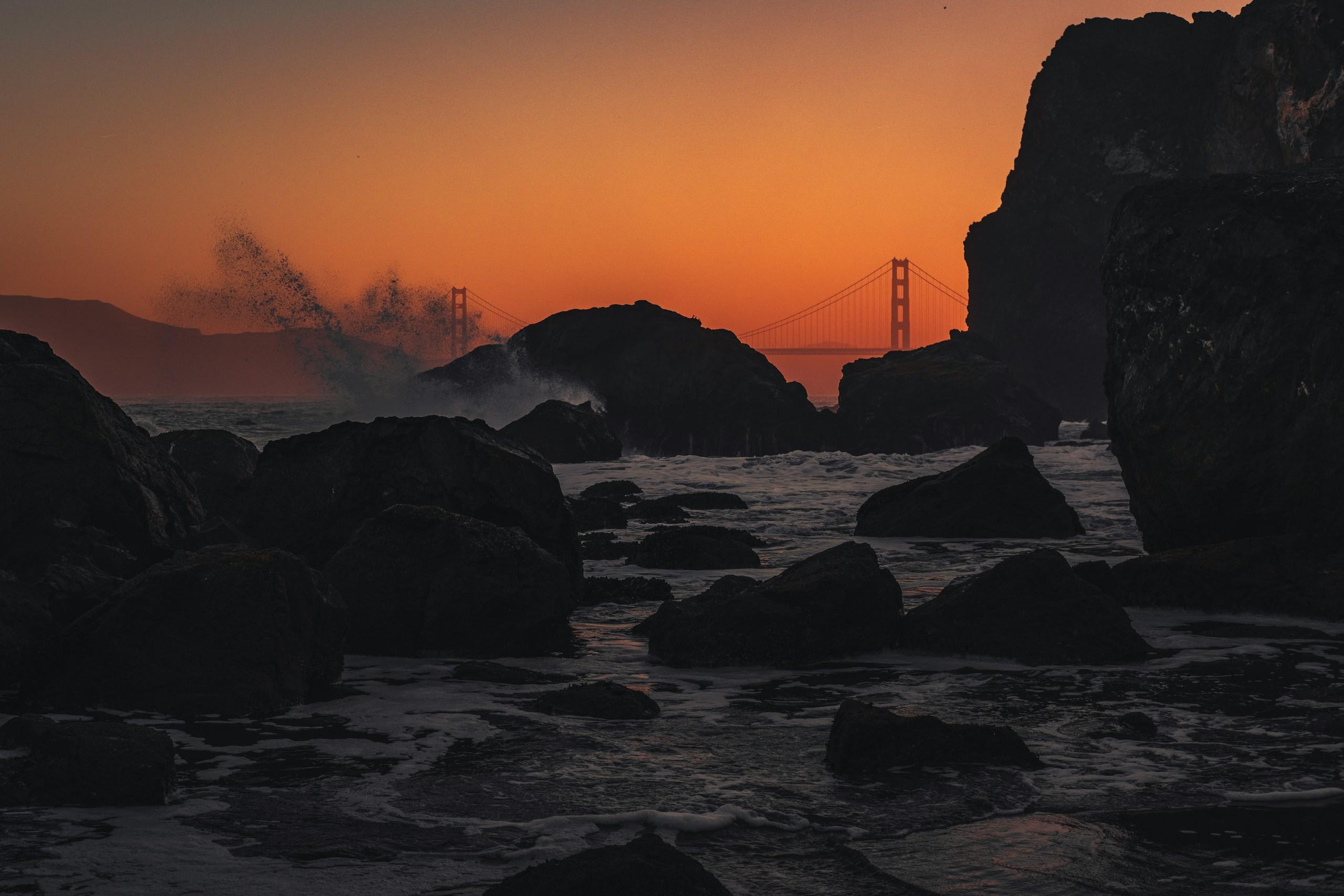 The Golden Gate Bridge in the distance behind dark rocky cliffs and crashing waves