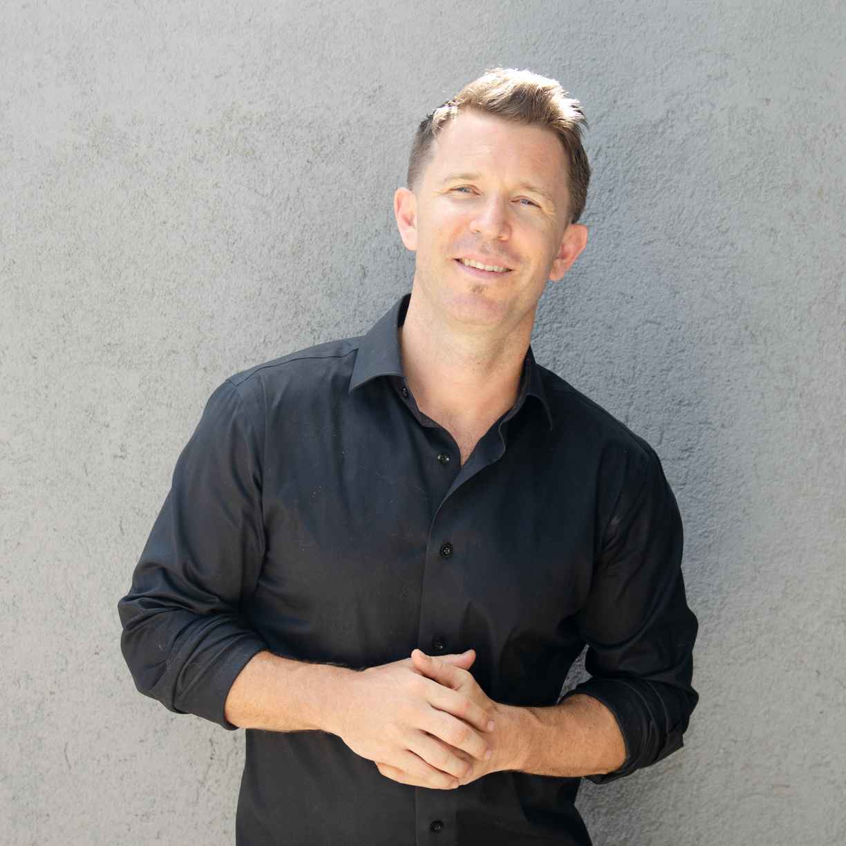 Damon smiling, wearing a black shirt with arms clasped in front of his chest against a gray concrete wall