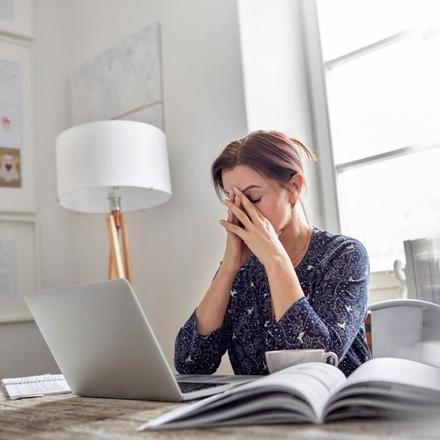 En kvinde sidder med lukkede øjne ved sit skrivebord. Hun tager sig til hovedet og udstråler stress. Hendes computer er åben og der er også en bog der ligger åben.