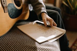 A picture of a notebook with lyrics on a couch being held by a person who si mostly out of the shot also holding an acoustic guitar