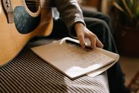 A person holding an acoustic guitar on their lap. In their had is a pen and notebook with lyrics