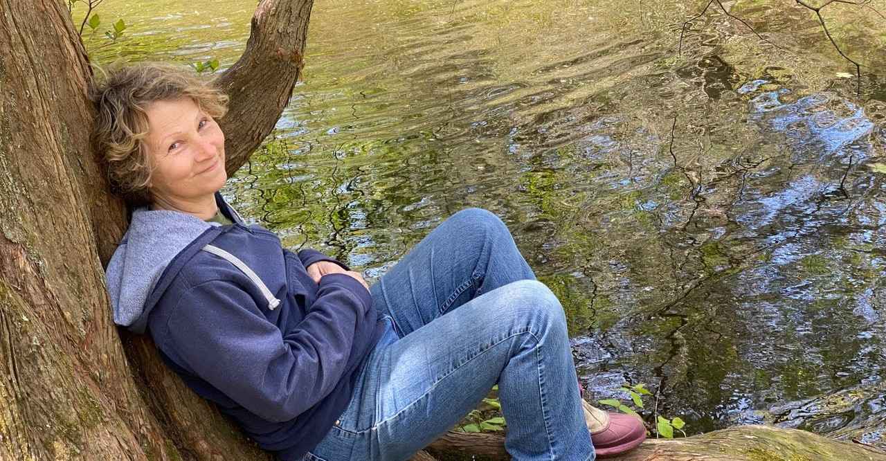 Nadine Mazzola, Certified Forest Therapy Guide sitting by water and leaning against a tree